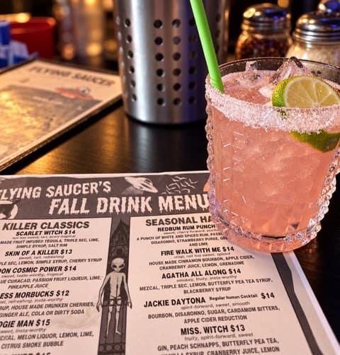 A pink cocktail with a sugared rim, a lime wheel and a green straw on a dark bar top, beside Flying Saucer Pizza Company's Fall Drink Menu listing Killer Classics and Seasonal Haunts cocktails