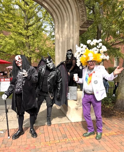 Three performers in black costumes and pale masks — one horned with leather bat wings, one in a long black wig with cracked white face paint holding a cane, one in goggles seated on a stone ledge behind a hand-lettered "Gargoyles of Salem" sign — standing under a carved stone arch beside a man in a paint-splattered white blazer, purple trousers and a tall yellow and white paper flower headdress