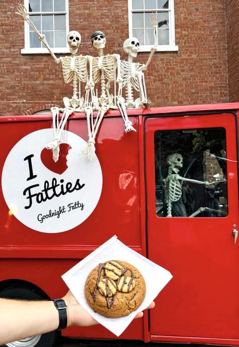 A hand holding a chocolate chip cookie drizzled with caramel on a white paper square, in front of Goodnight Fatty's red truck with its I Love Fatties sign, three skeletons sitting on the roof and another in the driver's seat