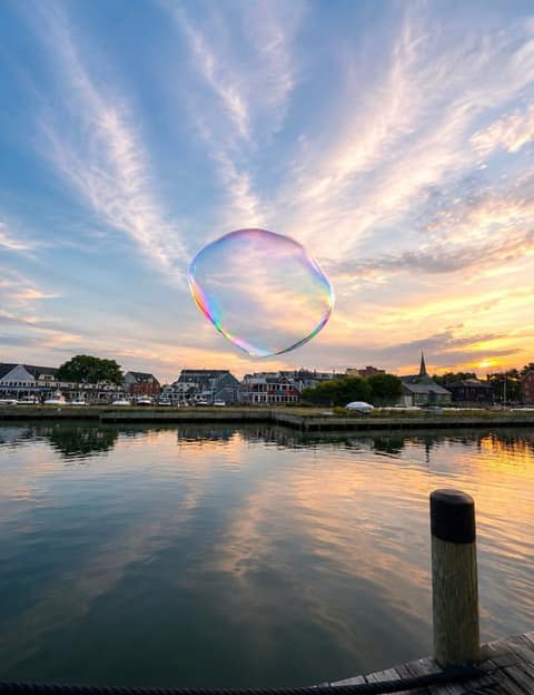 A giant iridescent soap bubble drifting over Salem Harbor at sunset, the waterfront skyline mirrored in calm water