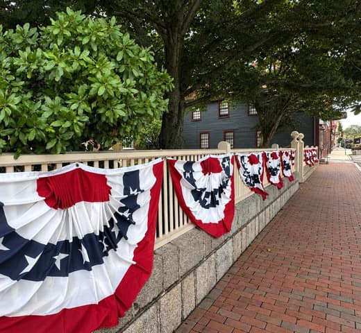 Red-white-and-blue bunting draped along a white fence on a brick sidewalk in Salem, with a historic dark clapboard building behind
