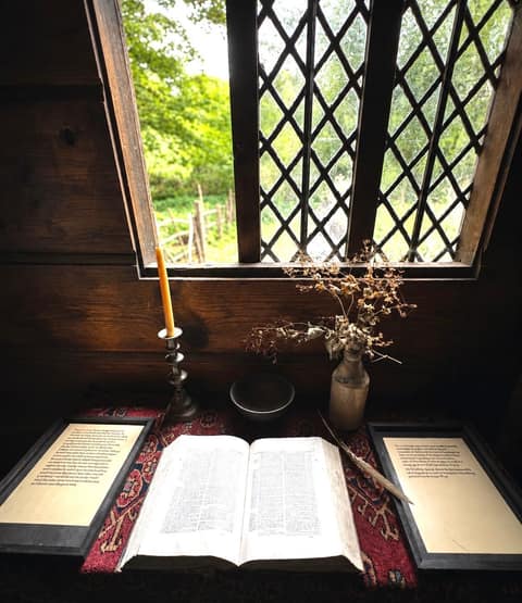 An open book, a candlestick and a quill pen on a table beside a diamond-paned casement window looking onto a garden