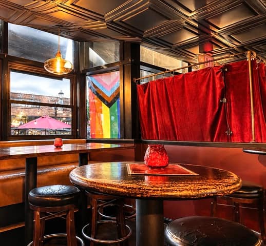 A corner booth and round bar table under a pressed-tin ceiling at All Souls Lounge, with a Progress Pride flag in the window and the street visible outside