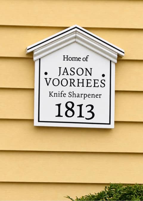 A white peaked house plaque in the style of Salem’s historic home markers, mounted on yellow clapboard siding, reading "Home of JASON VOORHEES, Knife Sharpener, 1813"