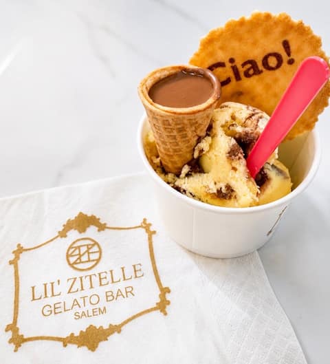 A white cup of swirled gelato topped with a waffle mini-cone full of chocolate and a round wafer stamped “Ciao!”, a pink spoon alongside, resting on a napkin printed with the Lil’ Zitelle Gelato Bar Salem logo