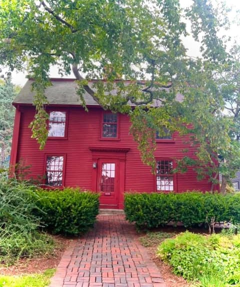 A dark red two-story colonial house with white-trimmed windows and a red front door, a brick walkway leading to it between clipped hedges, framed by a large tree.