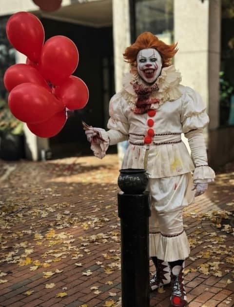 A street performer costumed as Pennywise the clown in a ruffled white suit with red pompoms, holding a bunch of red balloons beside a black bollard on a brick pedestrian street scattered with fallen leaves