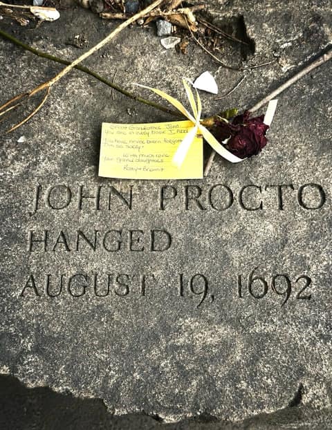 The John Proctor memorial bench at the Salem Witch Trials Memorial, inscribed 'hanged August 19, 1692', with a rose and a handwritten note left by a visitor