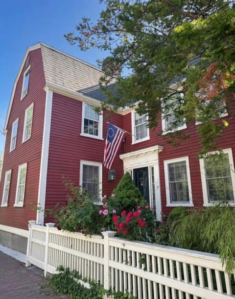 A deep-red gambrel-roofed colonial house with white trim, a 13-star flag, and a rose garden behind a white picket fence in Salem