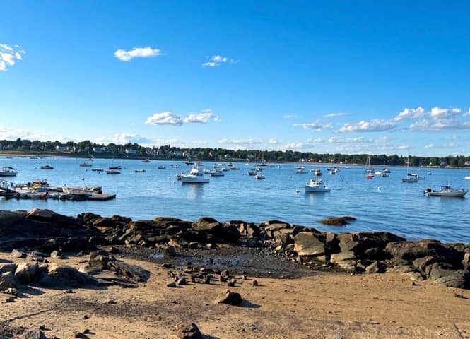 Recreational boats moored in Salem Harbor seen from a sandy beach with rocky outcrops, houses and a church steeple on the far shore, Salem MA