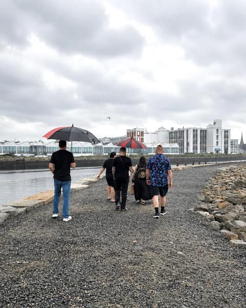 A group of visitors with red-and-black umbrellas walking the rocky causeway along Salem Harbor toward the Pickering Wharf waterfront, Salem MA
