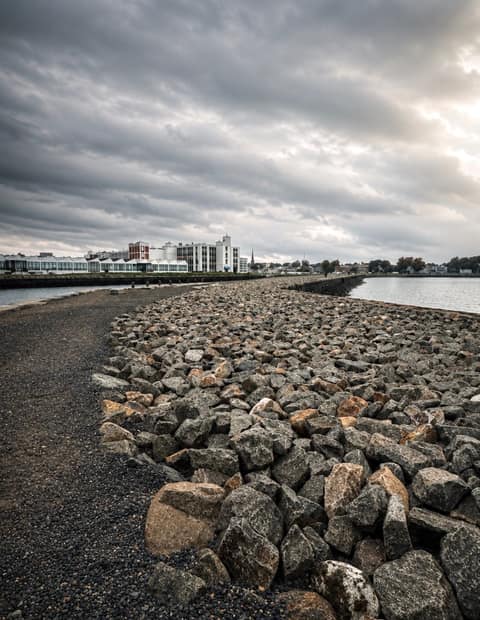Granite riprap breakwater curving along Salem Harbor toward the Pickering Wharf waterfront under a heavy cloudy sky, Salem MA