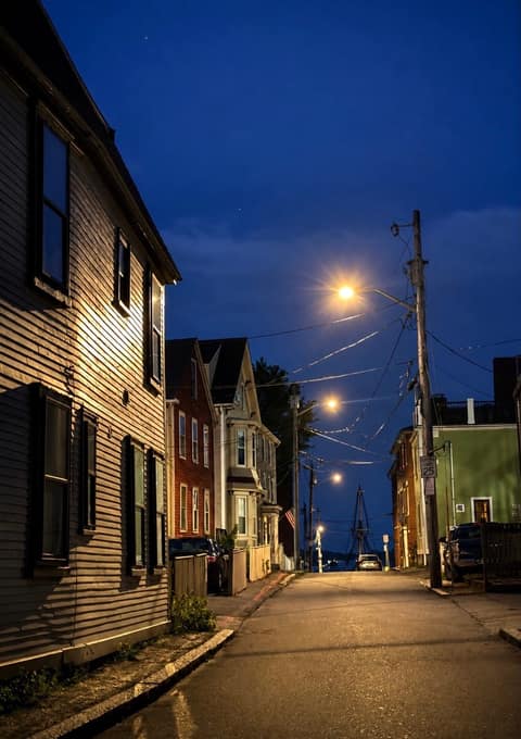 A quiet residential Salem street of clapboard houses sloping toward the harbor at blue hour, a tall-ship mast in the distance