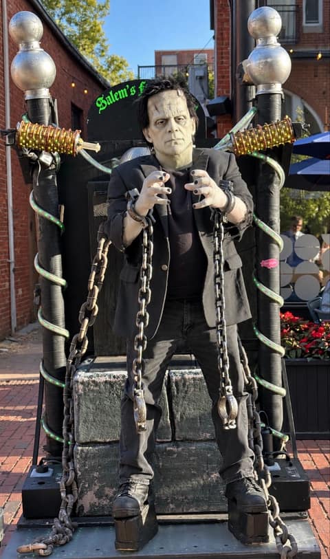 A street performer costumed as Frankenstein's monster sits chained to a heavy wooden throne flanked by two tall lamp posts wound with green cable, under a banner reading Salem's Frankenstein