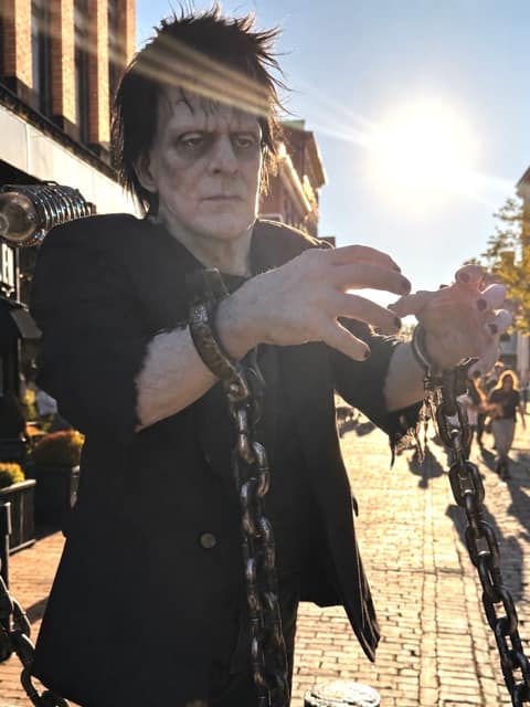 A street performer in full Frankenstein's monster makeup and a dark suit reaches both chained hands toward the camera on a sunlit brick pedestrian street
