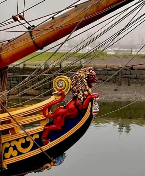 The bow of a tall ship in fog, with a red and gold carved lion figurehead beneath the bowsprit and rigging, moored beside a stone wharf.