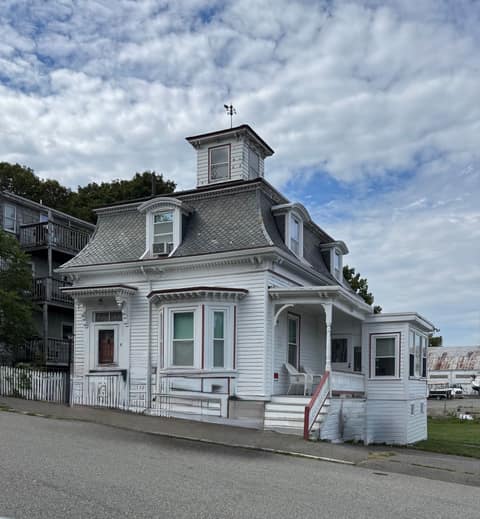 A white mansard-roofed house with a cupola and weathervane, seen from the street with a boatyard behind it