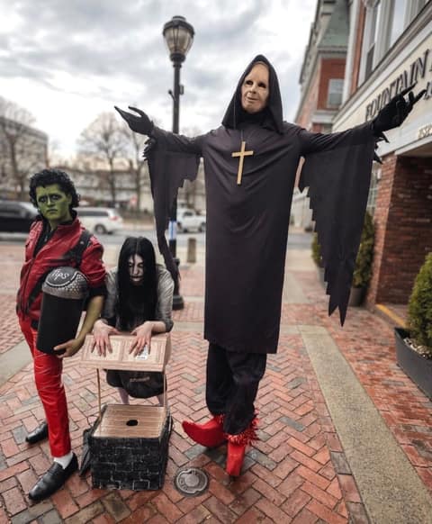 Three street performers on a brick sidewalk: one in green zombie makeup and a red zippered jacket holding a tips bucket, one costumed as Samara from The Ring with long black hair crouched behind a cardboard well prop, and a very tall figure in a black hooded robe, pale mask, wooden cross and red boots with arms spread wide