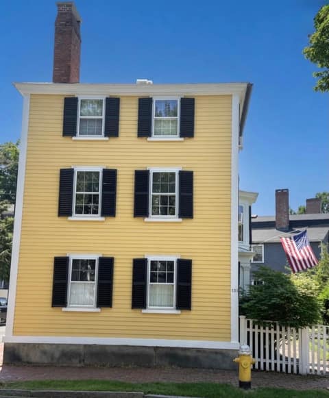 A three-story yellow Federal-style clapboard house with black shutters and a white picket fence under a blue sky in Salem MA