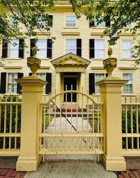 A three-storey yellow clapboard Federal-style house with black shutters and a pedimented doorway, seen through a matching yellow iron fence and arched gate with carved stone urns on the gateposts, framed by overhanging green branches