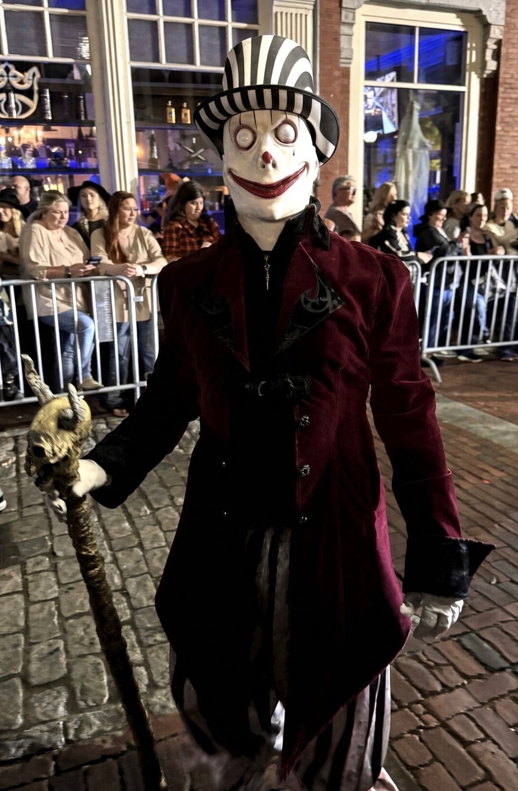 A figure in a smiling white porcelain-style mask, black-and-white striped top hat and burgundy velvet tailcoat, holding a twisted cane, walking past the barricades at the Grand Parade