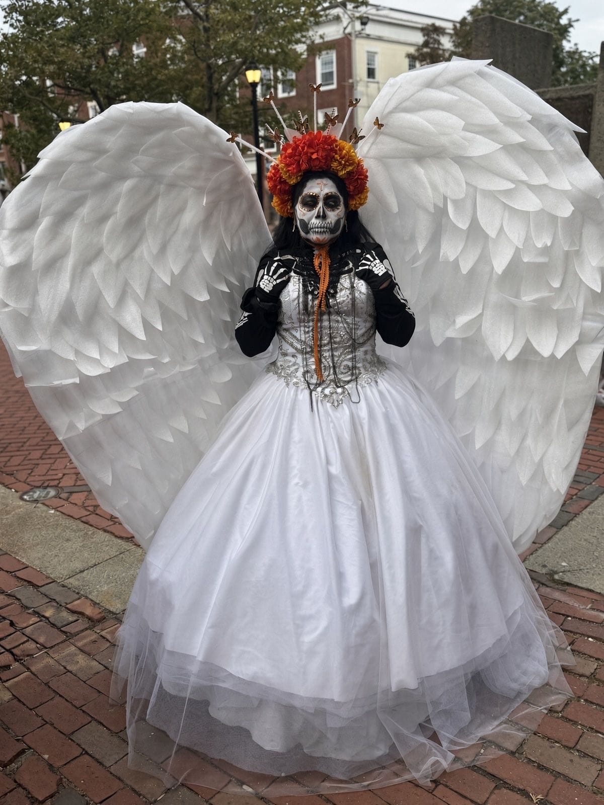 A woman in Day of the Dead skull makeup with a crown of orange marigolds, a white ball gown and enormous white feathered wings, posing on the brick street during the Grand Parade