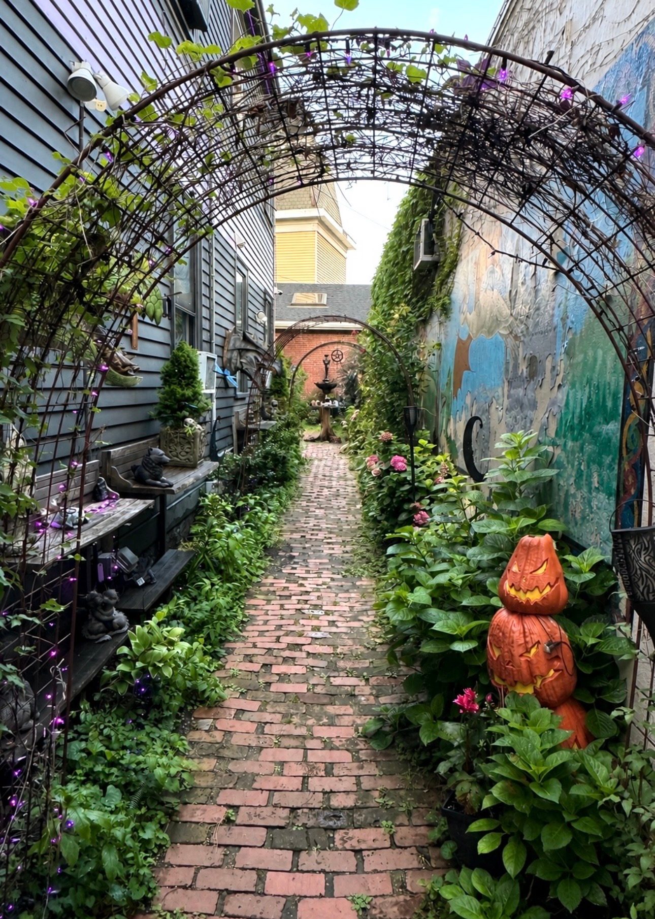 A narrow brick garden path under a vine-covered iron arch with greenery, a stacked jack-o-lantern totem, and a painted mural in Salem