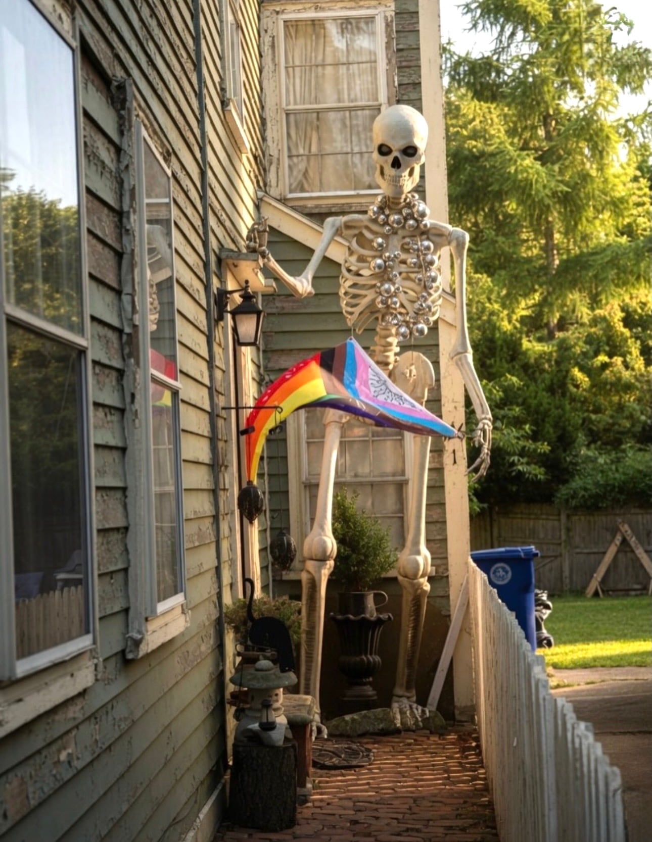 A twelve-foot skeleton decorated with pearls and holding a Progress Pride flag in the side yard of a weathered clapboard house in Salem MA