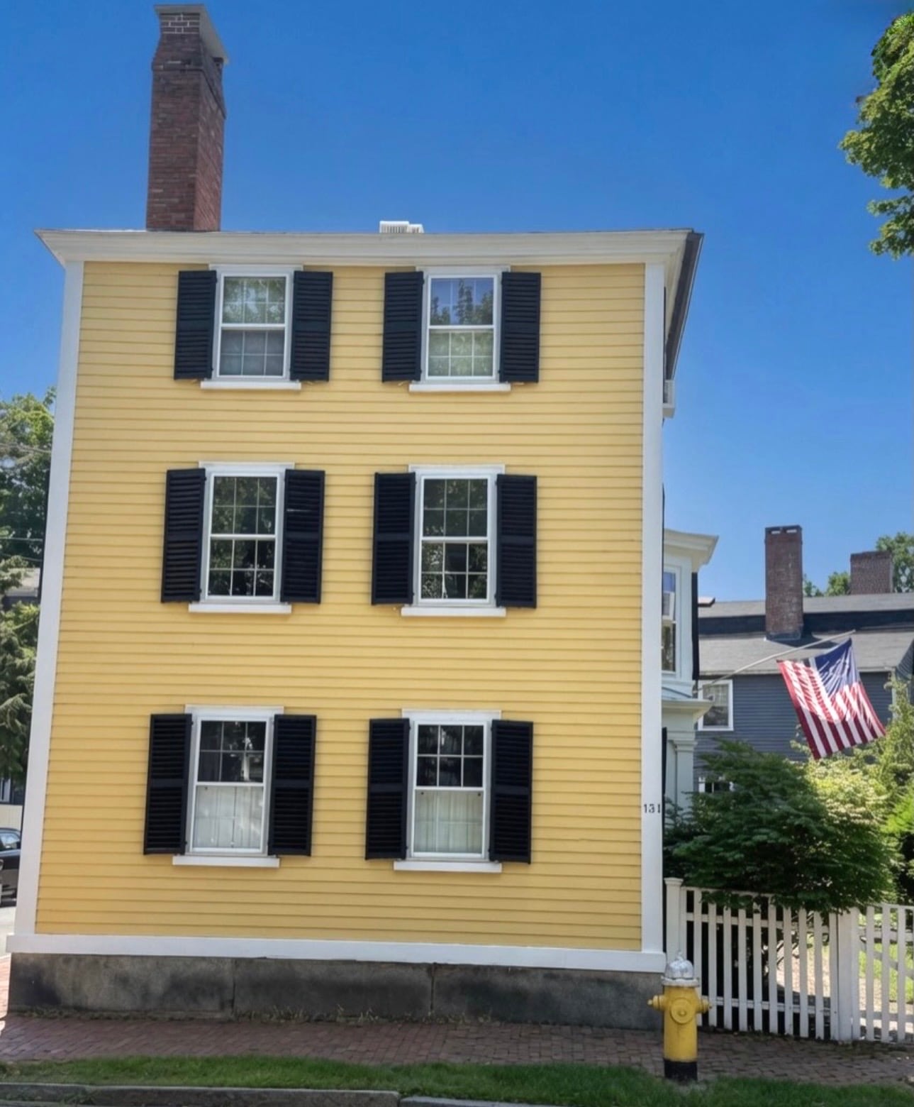 A three-story yellow Federal-style clapboard house with black shutters and a white picket fence under a blue sky in Salem MA