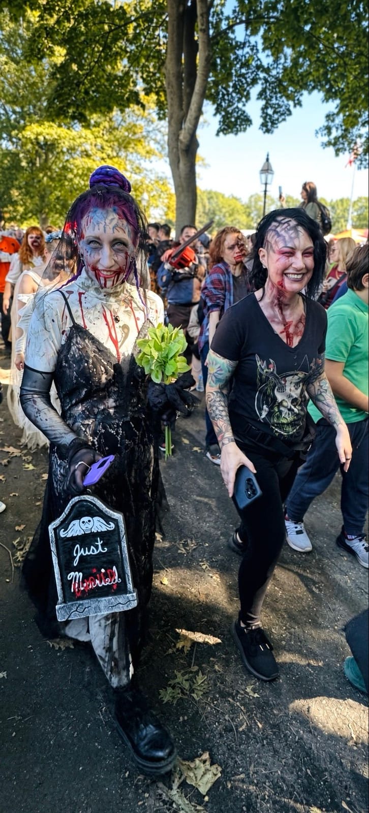 A zombie bride in a shredded black dress with blood down her chin, holding green flowers and a small tombstone sign reading Just Married, walking with a smiling zombie friend at the Salem Zombie Walk