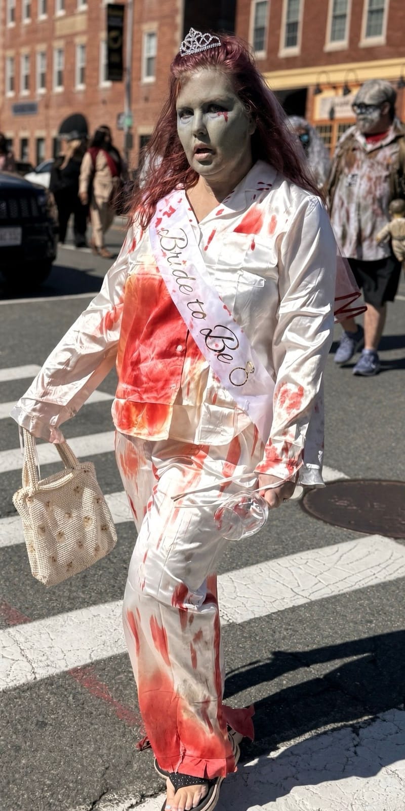A zombie in blood-spattered white satin pajamas, a tiara and a Bride to Be sash, carrying a beaded purse across a crosswalk with other zombies behind her